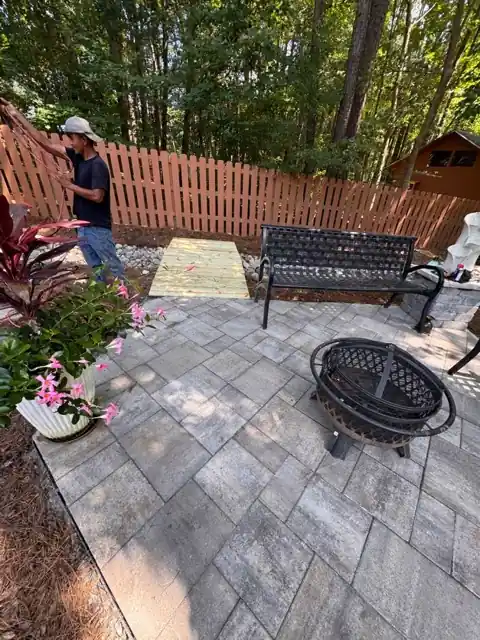 Grey paver patio with a fire pit, a wood bench and potted flowers under mature trees