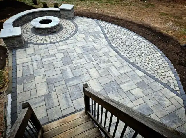 Large-format paver patio with a curved cobblestone accent band and a round fire pit, stepping down from a wood deck