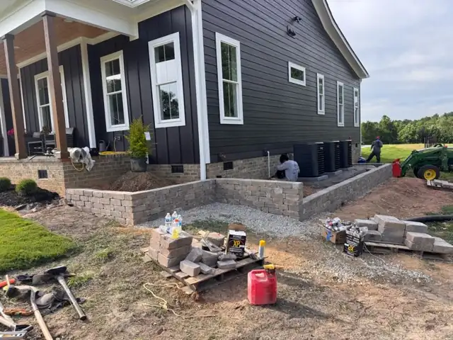 Block retaining wall being built around the base of a porch, with blocks and gravel staged nearby