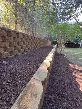 Long block retaining wall with a freshly mulched bed and young trees along a fence line