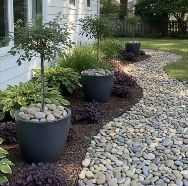River-rock bed with large dark planters, small trees and hostas beside a house